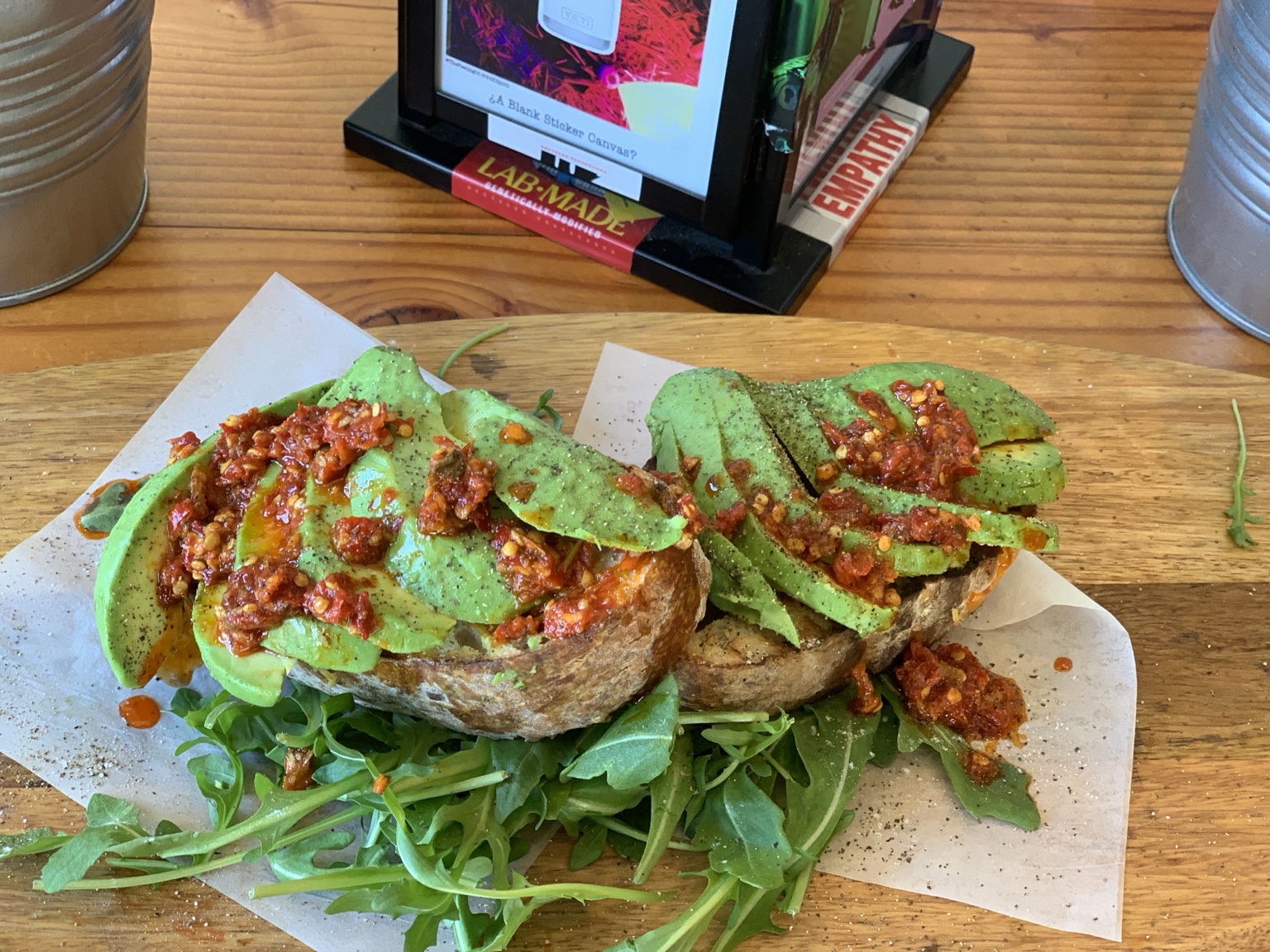 Two avocado toasts on sourdough, topped with red Calabrian chili peppers and served over arugula on a wooden board