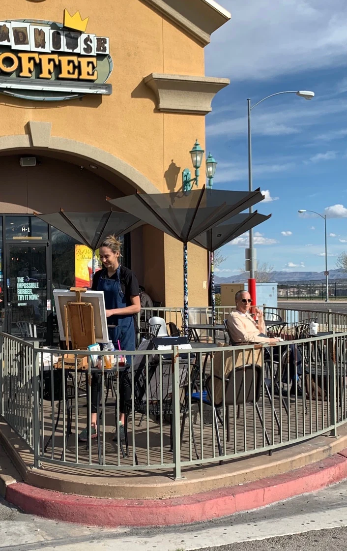 A woman paints on the patio while a man sits to her right beneath shade canopies