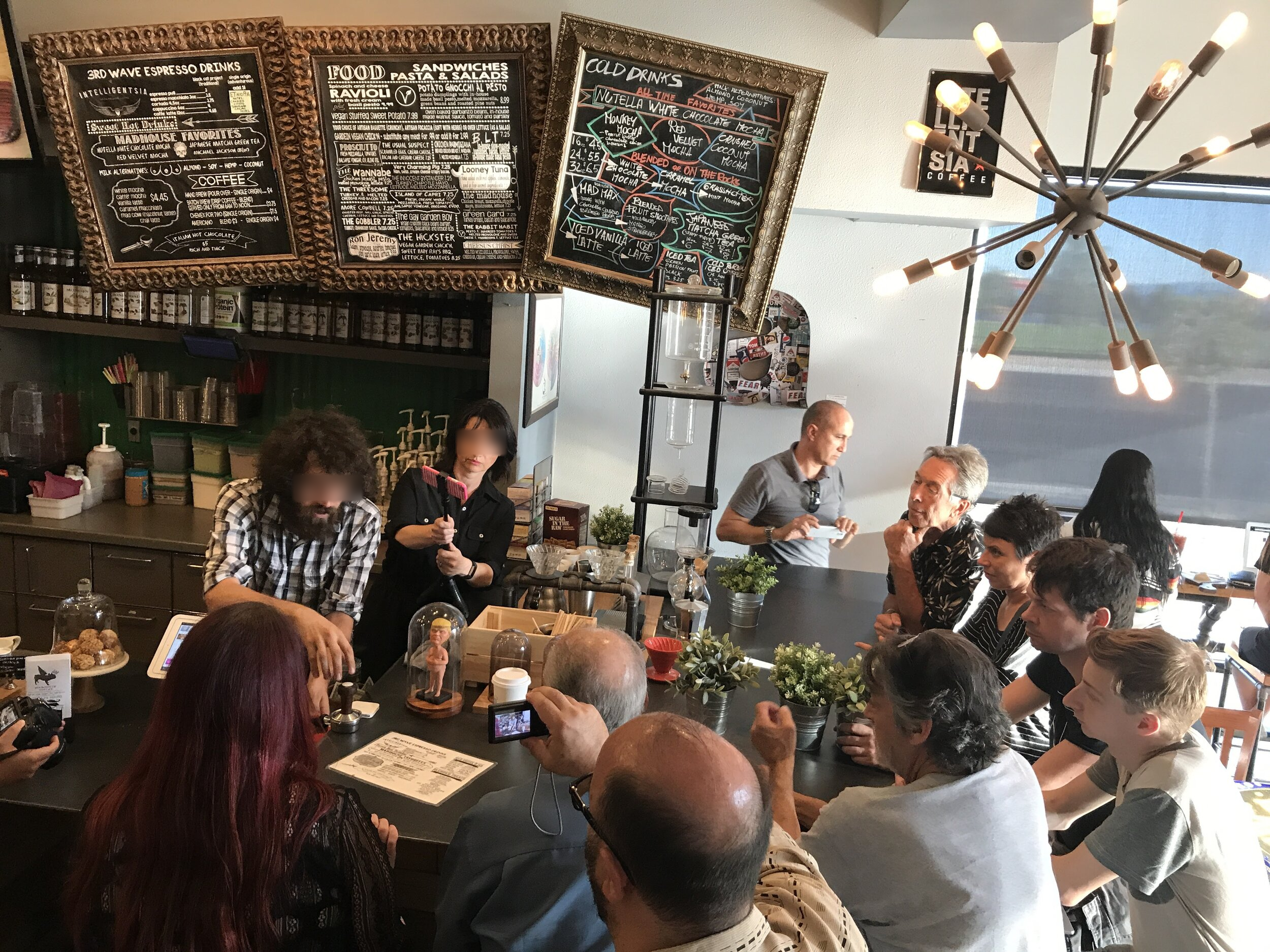 People gathered around tables and the counter beneath chalkboard menus and a branching light fixture