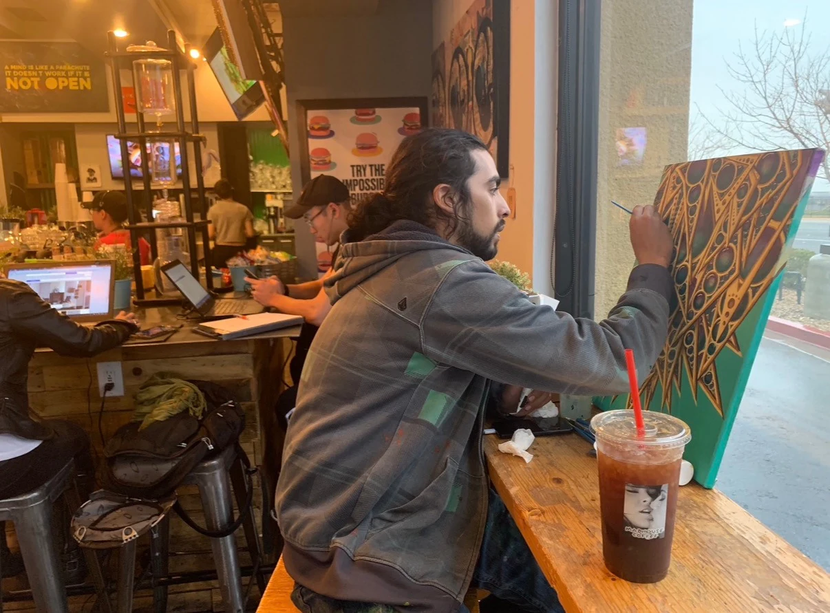 An artist paints by the window, with people using laptops at a shared table behind him
